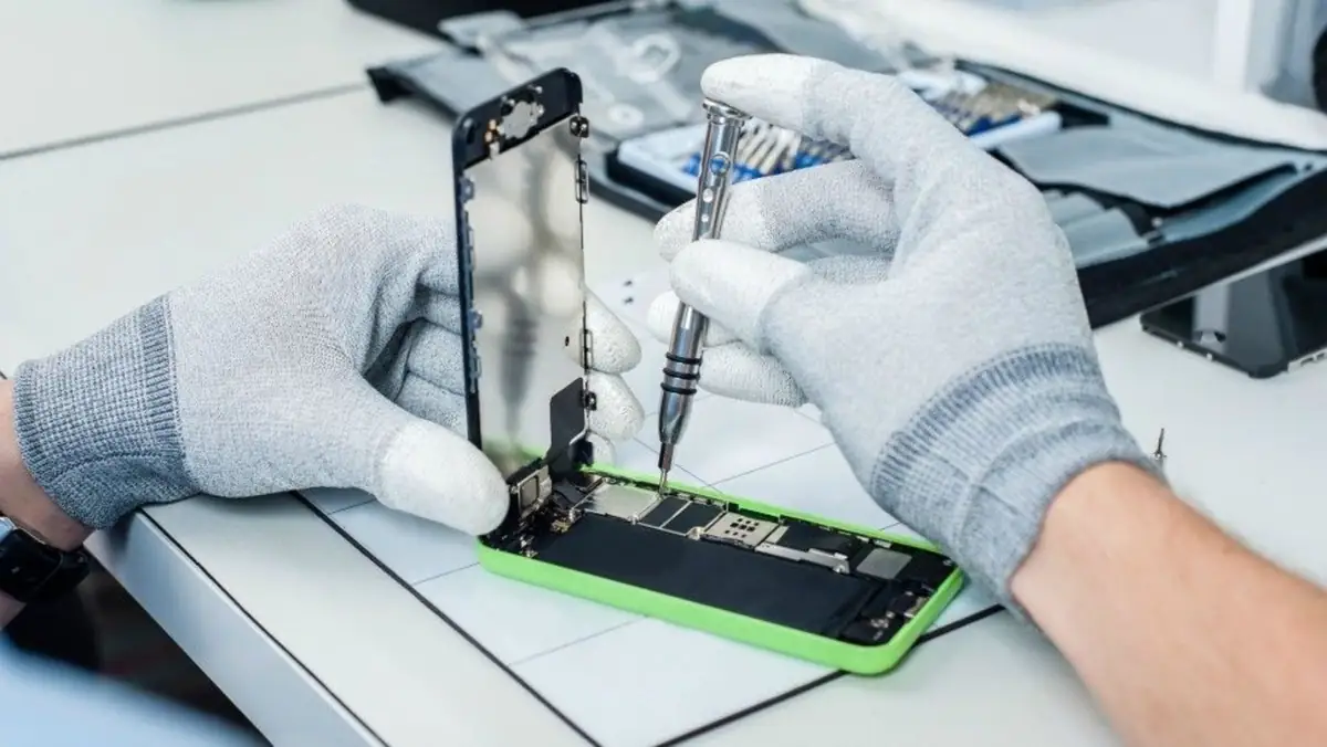 Mãos de luva desparafusam a parte de baixo de um iPhone 5c verde aberto, onde fica o conector.