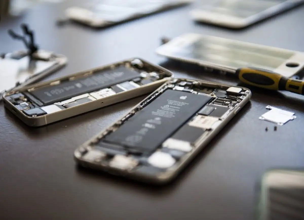 Dois iPhones abertos sobre a bancada, com bateria e placa à mostra e uma tela solta ao fundo.