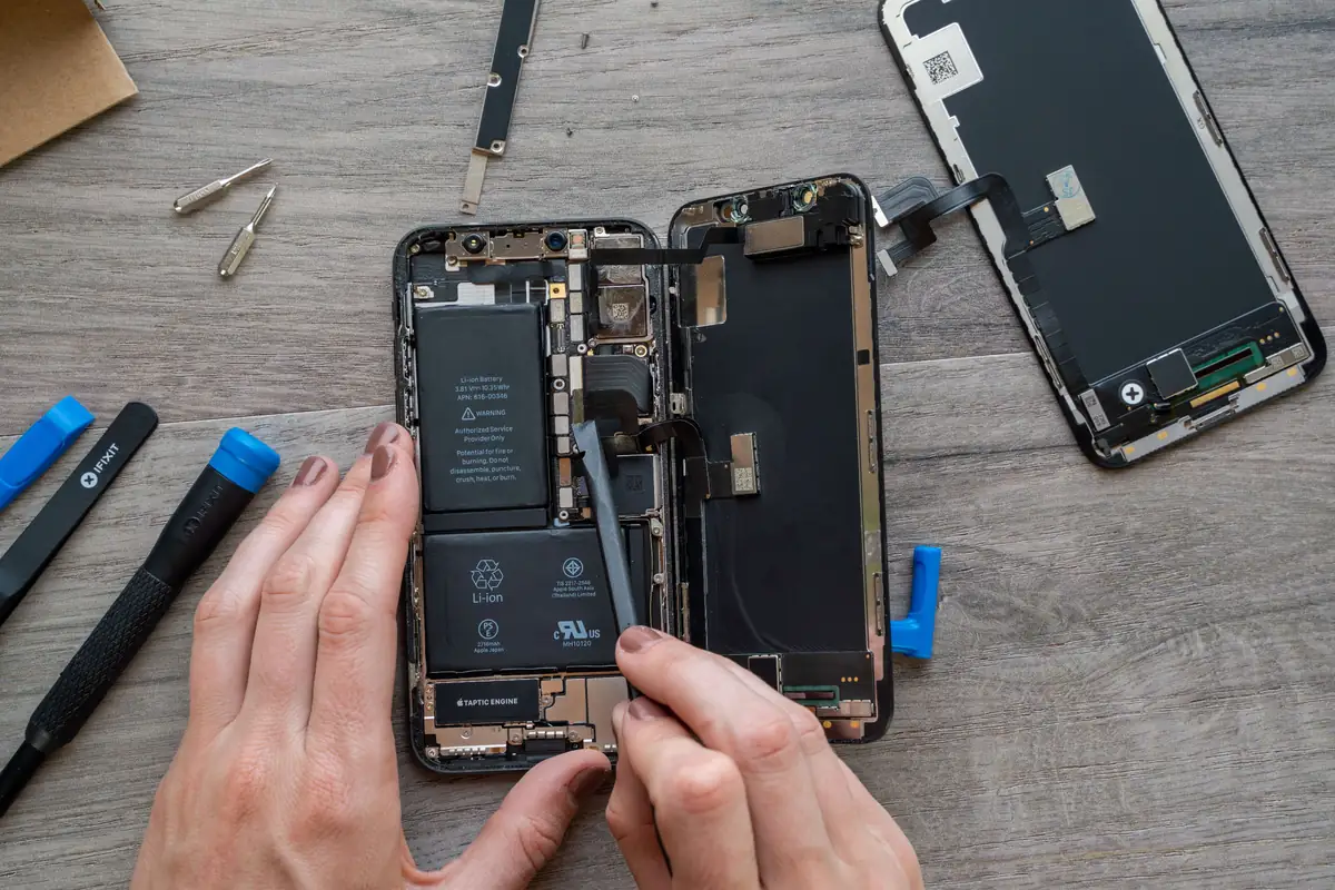 Mãos com ferramentas de abertura trabalham num iPhone aberto, com a tela solta ao lado.